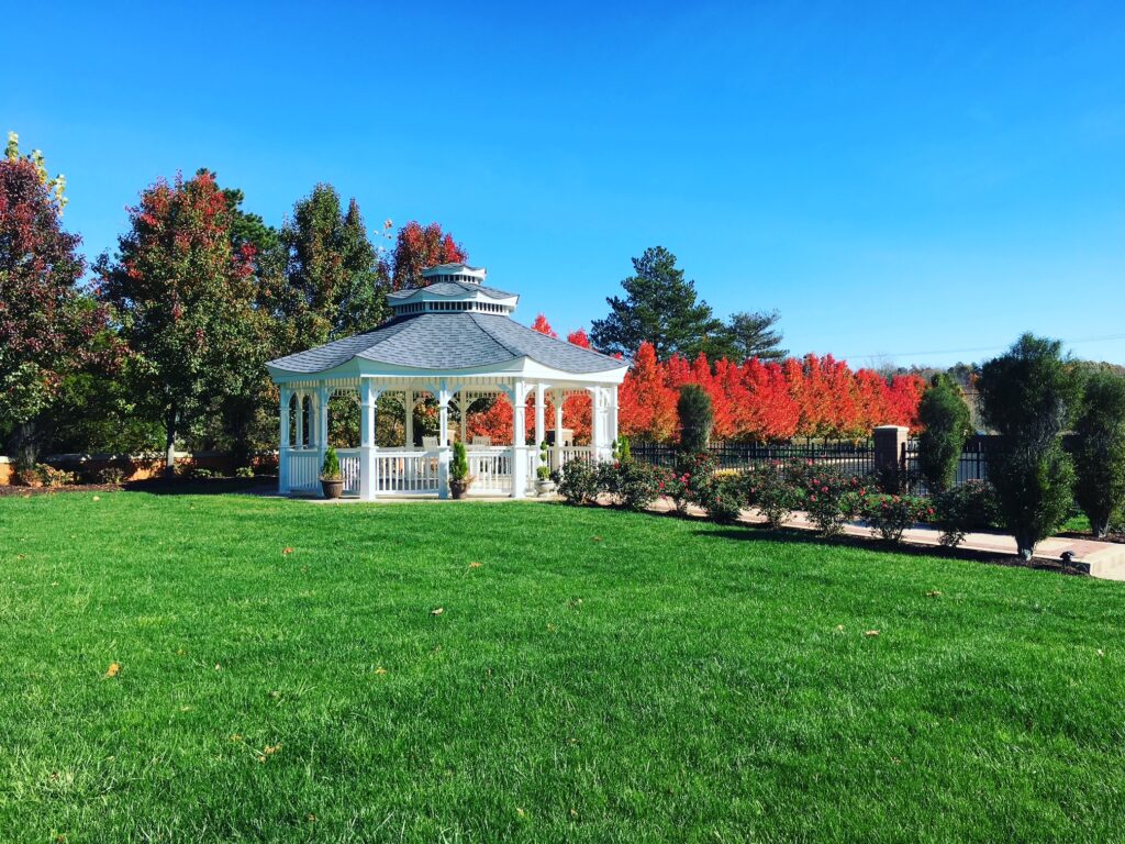 White gazebo surrounded by vibrant red and green trees on a lush lawn, showcasing a picturesque outdoor space ideal for gatherings