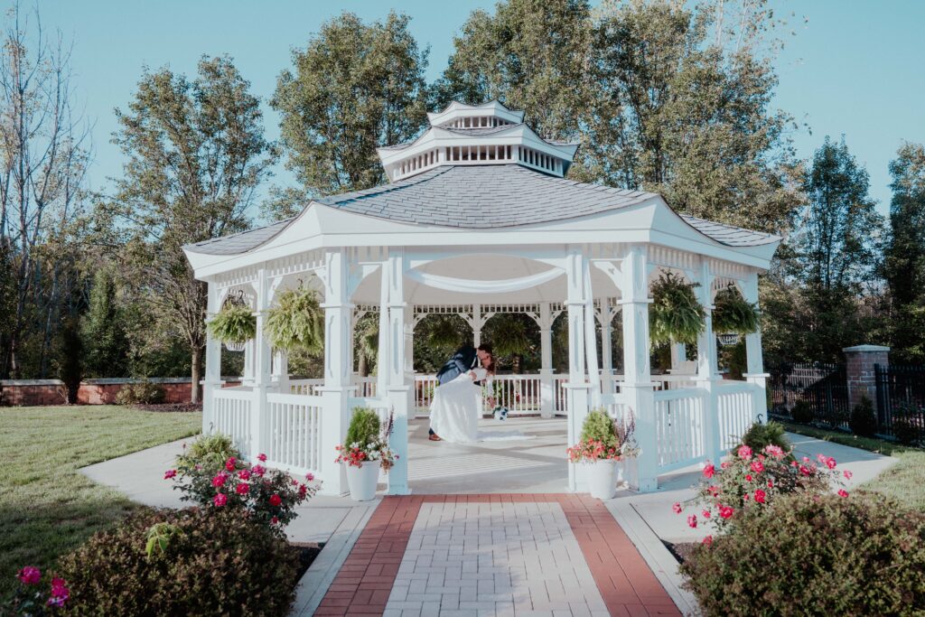 A bride and groom embrace in a white gazebo surrounded by greenery and colorful flowers