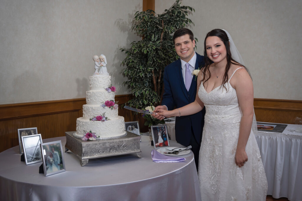A bride in a white gown and groom in a navy suit smile as they cut a three-tier wedding cake adorned with flowers