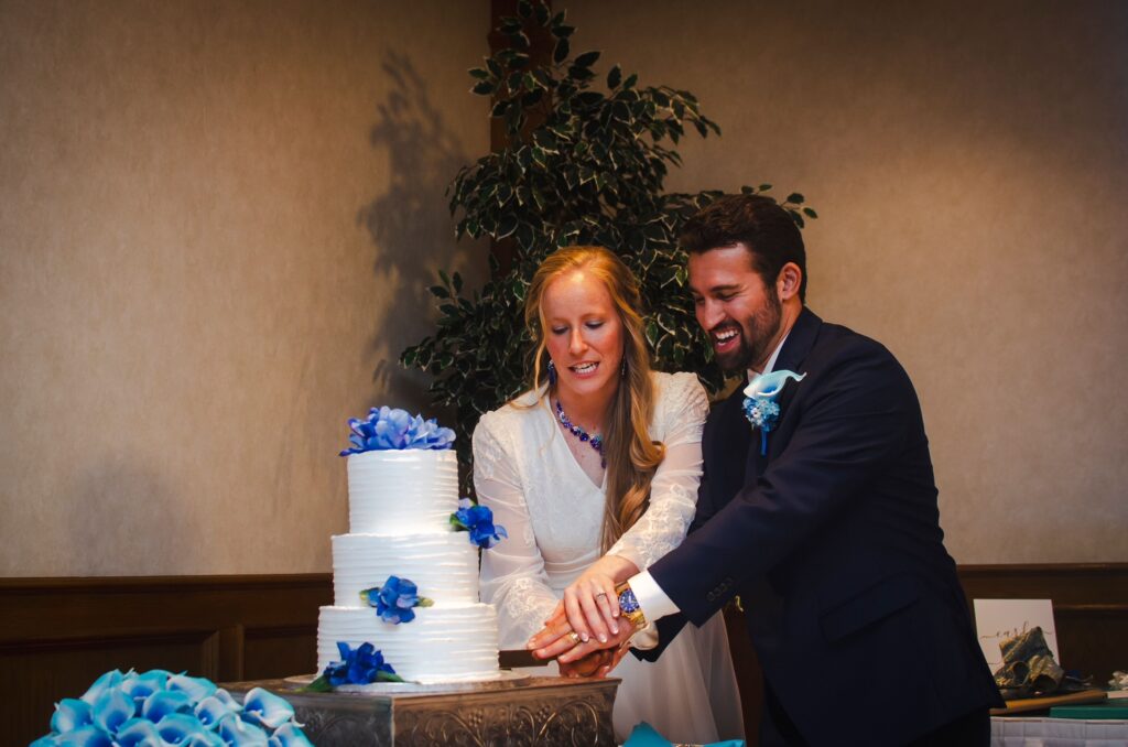 Bridal couple joyfully cutting a three-tiered wedding cake adorned with blue flowers,